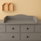 Eine graue Kommode mit sechs Schubladen und schwarzen Knöpfen wird von dem Wickelaufsatz grau passend für 160 cm breite HEMNES Kommode mit Blende gekrönt. Darüber ist an der beigen Wand ein KraftKids-Holzregal in Form von zwei Hausdächern angebracht.