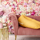 Ein rosafarbenes Samtsofa mit goldenen Beinen hält das KraftKids qualitäts Stillkissen Leinen gelb senfgelb, perfekt zum Schlafen und Stillen, neben einem runden goldenen Beistelltisch mit pastellfarbenen Blumen und einer leuchtend bunten Rosenwand im Hintergrund.