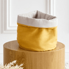 Ein KraftKids Körbchen rund Leinen gelb senfgelb mit hellgrau geripptem Innenfutter sitzt auf einem runden Holztisch an einer weißen Wand. Getrocknetes beiges Laub liegt davor-ideale Aufbewahrung für den Wickeltisch.