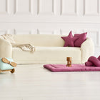 Vor einer weiß getäfelten Wand steht ein gemütliches weißes Sofa mit einer beigen Decke, zwei rosa Sternenkissen und Leinen Körbchen als Ablage. Davor liegt die KraftKids Krabbeldecke Leinen magenta dunkel, mit Holzklötzen, Kuscheltieren und einem hellblauen Spielzeugflugzeug.