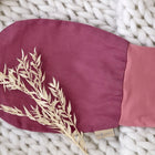 Ein magenta-dunkel gefärbter Strampelsack Herbst Winter Leinen von KraftKids mit einem kleinen beigen Anhänger ruht auf einer dicken weißen Strickdecke, die mit getrockneten beigen Blättern verziert ist.