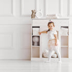 Ein junges Mädchen in einem Ballettkostüm lächelt neben einem weißen Aufbewahrungsschrank, der mit KraftKids Körbchen Musselin Zweige braun, gefalteten Baumwollhandtüchern und Dekoration in einem hellen, eleganten Raum mit weiß getäfelten Wänden gefüllt ist.