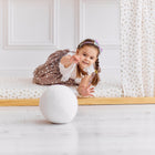 Ein junges Mädchen mit Zöpfen und einer Schleife liegt verspielt auf einem KraftKids Spannbettlaken Musselin Zweige braun und greift nach einem weißen Ball auf dem hellen Boden, der von gemusterten Vorhängen eingerahmt wird.