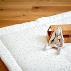 Die Krabbeldecke Musselin gelbe Zitronen von KraftKids, eine weiche weiße Spielmatte mit kleinen gelben Zitronenmustern, liegt mit einer Stoffhasenpuppe im Kleid und einem Spielzeug-Wohnmobil aus Holz auf einem Holzboden.