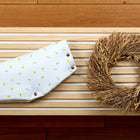 Der KraftKids Kinderwagenmuff Musselin gelbe Zitronen, ein handgenähter weißer Muff mit gelbem Zitronenmuster und Druckknöpfen, wird neben einem Weizenkranz auf hellen Holzlatten präsentiert.