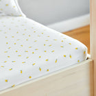 Eine Nahaufnahme eines Bettgestells aus hellem Holz mit den KraftKids Spannbettlaken Musselin gelbe Zitronen, die mit weißem Stoff mit kleinen gelben Zitronenmustern versehen sind. In dem hellen, sanft beleuchteten Raum sind ein passendes Kissen und ein Hartholzboden zu sehen.