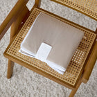 Die KraftKids Reisewickelunterlage Musselin beige liegt ordentlich auf einem Holzstuhl und eignet sich dank weißem Gurt ideal als Wickelauflage für unterwegs - perfekt für modernes Reisen mit Baby.