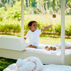 Ein kleines Kind in Weiß sitzt auf dem KraftKids Bettschutzgitter Rausfallschutz mit Stauraum, 60 cm breit, lächelt und wirft ein Holzspielzeug. Weiche Kissen, Decken und grünes Gras vervollständigen die friedliche Umgebung.