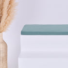 Eine hohe geflochtene Vase mit Pampasgras steht neben einer minimalistischen weißen Bank mit einem KraftKids GmbH Sitzauflage für Ikea TROFAST Musselin nilblau Kissen vor einem schlichten hellen Hintergrund.