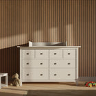 Ein weißer KraftKids Wickelaufsatz (gerade Front) passt auf eine 160 cm lange HEMNES Kommode, die an einer Holzlattenwand steht, neben einem kleinen weißen Stuhl, einem Plüschbären und einem Spielzeugauto auf dem Boden in diesem sanft beleuchteten Raum.