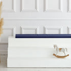 Ein minimalistisches Zimmer mit einer großen beigen Vase mit Pampasgras, einem weißen dreistufigen Podest mit dem KraftKids GmbH Sitzauflage für Ikea TROFAST Leinen blau dunkelblau Kissen und einem kleinen weißen Schaukelpferd. Im Hintergrund eine weiß getäfelte Wand.