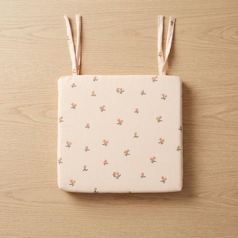 Das KraftKids Sitzkissen für Stühle aus Musselin ist ein quadratisches, hellrosafarbenes Sitzkissen mit zartem Blumenmuster und zwei Stoffbindebändern - ideal als Sitzkissen Esszimmer und hier auf einer hellen Holzfläche platziert.