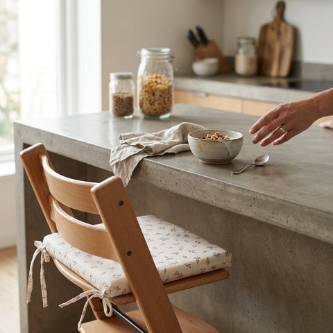 Eine Hand greift nach einem Müsli auf einer grauen Theke, mit Gläsern im Hintergrund und einem KraftKids Sitzkissen für Stühle Musselin 28x28 cm mit einem gemusterten Bezug auf dem Holzstuhl im Vordergrund.