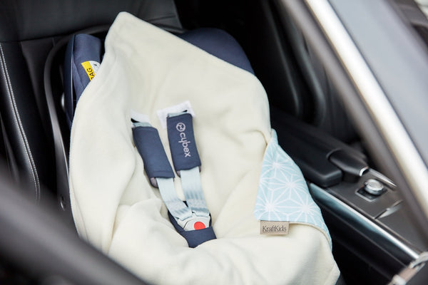 Eine KraftKids "Decke für Babyschale Winter weiße Diamante auf Pastell Blau" bedeckt einen Babyautositz auf der Rückbank eines Fahrzeugs. Die blau gemusterte Ecke, der Gurt und das Cybex-Etikett sind sichtbar, aber der Sitz ist leer.
