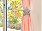 Eine Nahaufnahme einer rosafarbenen Gardine mit dem KraftKids "Gardinen Raffhalter weiße Punkte auf Grau", sternförmig mit weißen Punkten auf Grau, ist neben einem Fenster mit Blick auf Herbstbäume mit gelben und orangefarbenen Blättern zu sehen.