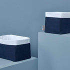 Zwei KraftKids Körbchen Musselin dunkelblau - einer rechteckig, einer quadratisch - mit dunkelblauem Rand und weißem Deckel sind perfekt fürs Kinderzimmer. Abgebildet auf hellblauen Sockeln.