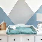 Auf einer weißen Kommode mit schwarzen Knöpfen stehen eine KraftKids Wickelauflage Musselin nilblau, zwei Spielzeugschafe, ein weicher blauer Hase, eine Lampe und hausförmige Regale. Die Wand dahinter zeigt ein auffälliges geometrisches Muster in Blau und Weiß.