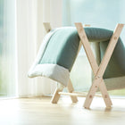 Die KraftKids Krabbeldecke Musselin mint, eine weiche grün-weiße Babydecke, ruht auf einem hellen Holzständer in der Nähe eines großen Fensters, daneben steht ein geflochtener Korb mit rosa Bommeln - perfekt, um gemütliche Momente zu schaffen.