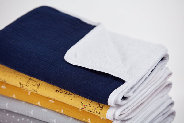 Ein Stapel gefalteter Babydecken in Marineblau, Weiß, Gelb mit Hühneraufdruck und Grau mit kleinen weißen Mustern liegt ordentlich neben der KraftKids Wickelunterlage Musselin dunkelblau auf einer weißen Fläche.