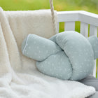 Ein KraftKids Wickeltischumrandung Musselin mint Pusteblumen in Hellgrün mit weißem Pusteblumenmuster liegt auf einer weichen cremefarbenen Decke über einem weißen Kinderbett, akzentuiert durch ein graues Kissen und unscharfe Grünpflanzen im Hintergrund.