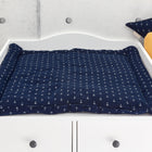 Ein KraftKids Wickelauflage Musselin dunkelblauer Anker mit weißem Ankermuster liegt auf einer weißen Kommode, neben einer mit Wattestäbchen gefüllten Stoffbox, einem marineblauen Sternkissen und einem gelben apfelförmigen Kissen.