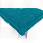 Eine KraftKids Wickelauflage Musselin in Petrol mit goldenen Punkten wird ordentlich gefaltet und auf einer weißen, geometrischen Fläche präsentiert.