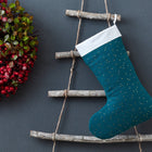 Ein festlicher Kranz mit roten Beeren hängt an einer grauen Wand neben einer rustikalen Leiter, auf der die KraftKids Weihnachtssocke Musselin goldene Punkte auf Petrol, ein blauer Weihnachtsstrumpf mit goldenen Punkten und weißem Bündchen, hängt.