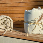 Zwei KraftKids Bettumrandung Nestchen Leinen in leicht grünem Weiß sind auf einer hölzernen Unterlage gerollt; eines ist mit Bindfaden und getrockneten Gräsern verschnürt, das andere lose mit sichtbaren Stoffbändern gerollt.