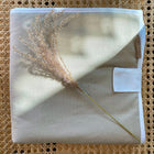 Die KraftKids Reisewickelunterlage Leinen Natur Braun, eine beigefarbene Leinen-Wickelunterlage mit weißem Rand und Anhänger, liegt auf geflochtenem Rattan, wie getrocknetes Gras das Sonnenlicht einfängt - ideal als stilvolle Wickelauflage für unterwegs oder platzsparende Wickelunterlage.