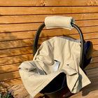 Eine schwarze Babyschale mit beigem Stoff und Kopfstütze steht auf einem Holzdeck, das von grünen Pflanzen akzentuiert wird, gekrönt von der KraftKids Babydecke für Babyschale Sommer Leinen Natur Braun.