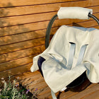 Eine KraftKids Babydecke für Babyschale Sommer Leinen leicht grünes Weiß bedeckt einen Babyautositz mit hellbeigem Bezug und grauen Gurten auf einem Holzdeck an einer Wand, mit Lavendelpflanzen in der Ecke.