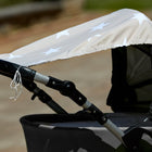 Nahaufnahme eines grauen Kinderwagens mit einem KraftKids Sonnensegel große weiße Sterne auf beigem Baumwollverdeck. Die Kanten haben ausgefranste Fäden, und der Hintergrund im Freien ist unscharf.