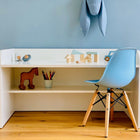 Der KraftKids Kinderschreibtisch weiß mit Zwischenboden steht auf einem Holzboden mit Holzspielzeug, Buntstiften in einer Tasse und einem blauen Stuhl vor einer blauen Wand, die mit einem ausgestopften Hasenkopf verziert ist.