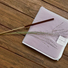 Ein gefaltetes Lavendeltuch, ein braunes Räucherstäbchen und getrocknetes Gras liegen auf Holz - perfekt neben der KraftKids Reisewickelunterlage Doppelkrepp Rosa für stilvolles, organisiertes Reisen.