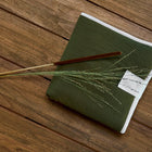 Die KraftKids Reisewickelunterlage Doppelkrepp Grün Herbstgrün ist auf Holz gefaltet und mit einem weißen Etikett versehen. Obenauf liegen braune und gelbe Stäbchen sowie getrocknetes Gras - perfekt für praktische Mütter, die viel unterwegs sind.