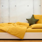 Ein minimalistisches Holzbett mit dem Bettwäscheset Doppelkrepp Gelb Senf von KraftKids - 100 % Baumwolle, mit passendem Kissen und grünem Sternkissen - sorgt vor einer hellen Betonwand für erholsamen Schlaf.