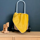 Ein kleines Holzpferdchen steht neben einem Weidenkorb mit weißem Griff, der mit der KraftKids Babydecke Cord Breitcord Gelb Senf drapiert ist, auf einem Holzschrank vor einer dunkelgrauen Wand.