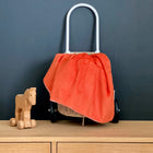 Über einem Weidenkorb auf Rädern mit weißem Griff ist eine KraftKids Babydecke Cord Breitcord Orange Reinorange drapiert, die auf einer Holzfläche neben einem kleinen Holzspielzeugpferd mit Rädern steht, alles vor einem schwarzen Hintergrund.