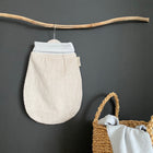 Ein KraftKids Strampelsack Frühling Sommer Cord Breitcord in Beige Taupe hängt an einem Holzast vor einer dunklen Wand. Ein geflochtener Korb mit Griffen darunter hält ein hellgraues Tuch.