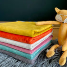 Ein Stapel bunter Decken liegt auf einer grauen, pelzigen Oberfläche neben einem orangefarbenen Fuchsplüsch mit geschlossenen Augen und einem geblümten Stirnband, alles auf der KraftKids Wickelunterlage Cord Breitcord Gelb Mustard.