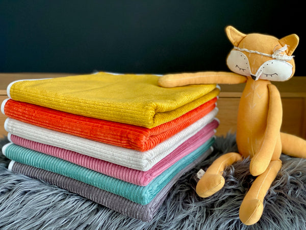 Ein Stapel bunter Decken liegt auf einer grauen, pelzigen Oberfläche neben einem orangefarbenen Fuchsplüsch mit geschlossenen Augen und einem geblümten Stirnband, alles auf der KraftKids Wickelunterlage Cord Breitcord Gelb Mustard.