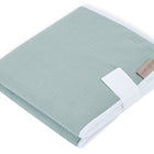 Eine sauber gefaltete KraftKids Reisewickelunterlage Musselin mint, mit weißem Rand und einem hellbraunen Etikett, gesichert mit einem weißen Stoffband, auf weißem Hintergrund.