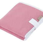Eine sauber gefaltete Reisewickelunterlage Musselin rosa von KraftKids aus zartrosa Musselin mit weißem Rand und einem kleinen braunen Anhänger auf weißem Hintergrund - ideal als platzsparende Wickelauflage für Unterwegs.