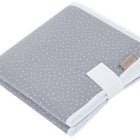 Die KraftKids Reisewickelunterlage Musselin grau Punkte ist eine sauber gefaltete graue Reisewickelunterlage mit weißen Punkten, weißem Rand, einem beigen Stoffanhänger in einer Ecke und einem weißen Streifen, der teilweise um die Unterlage gewickelt ist.