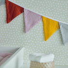 Eine Nahaufnahme einer gepunkteten Wand, die mit der KraftKids Wimpelkette Doppelkrepp Grau Gelb Ros Herbstrot dekoriert ist. Rote, rosa, gelbe und graue Dreiecke setzen einen fröhlichen Akzent über einem Teil eines Kinderbettes mit rosa Bettwäsche im Vordergrund.