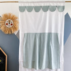 Ein KraftKids Puppentheater Kasperletheater aus Musselin mit goldenen Punkten auf Grün hängt vor einer Wand mit geometrischen Mustern. Links davon sind ein Strohlöwen-Deko und ein Teil einer beschrifteten schwarzen Tafel zu sehen.